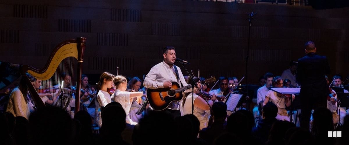 A guitarist and vocalist performs at a microphone stand with a full orchestra behind him in a concert hall, a harp visible on the left and a conductor on the right.
