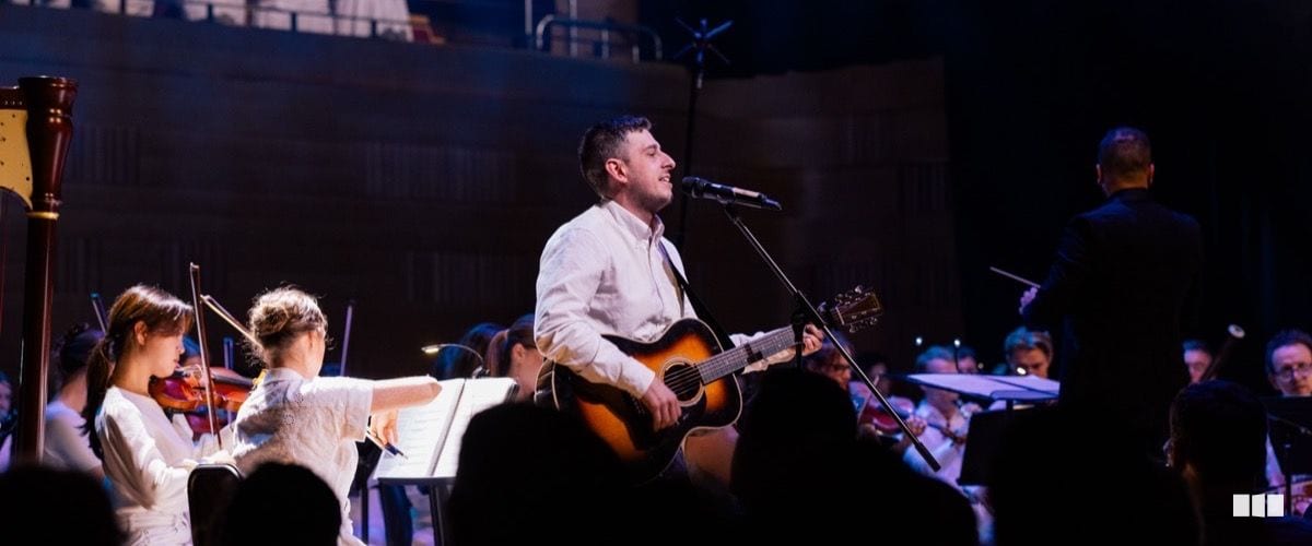 A male soloist plays acoustic guitar and sings into a microphone at centre stage, backed by a full orchestra including violin players in white.