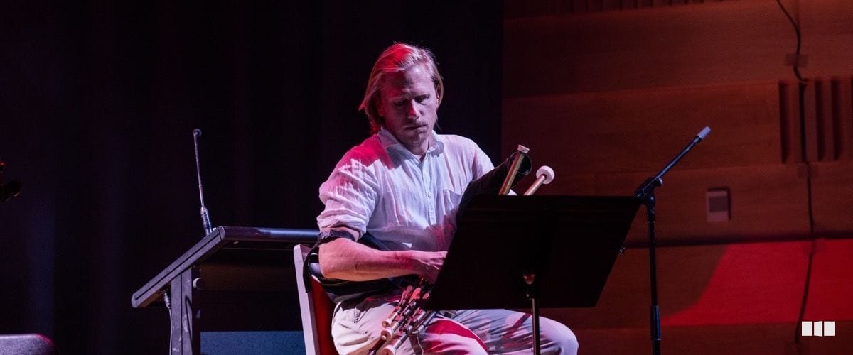 A male musician seated on stage reads from a music stand while holding a woodwind instrument under dramatic red stage lighting.