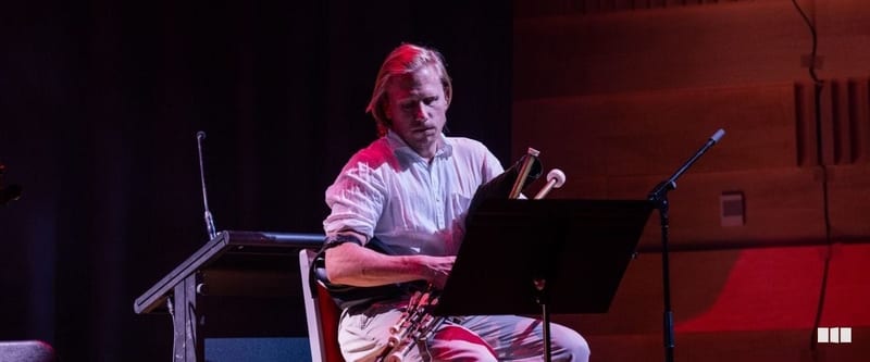 A male musician seated on stage reads from a music stand while holding a woodwind instrument under dramatic red stage lighting.