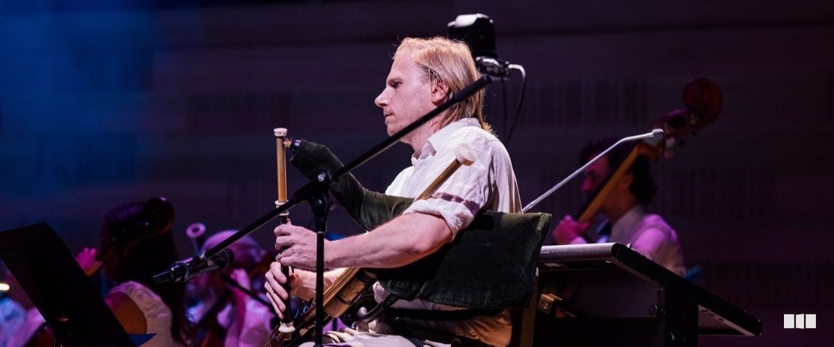 A blond male musician plays a traditional wind instrument while seated on stage with an orchestra ensemble behind him.