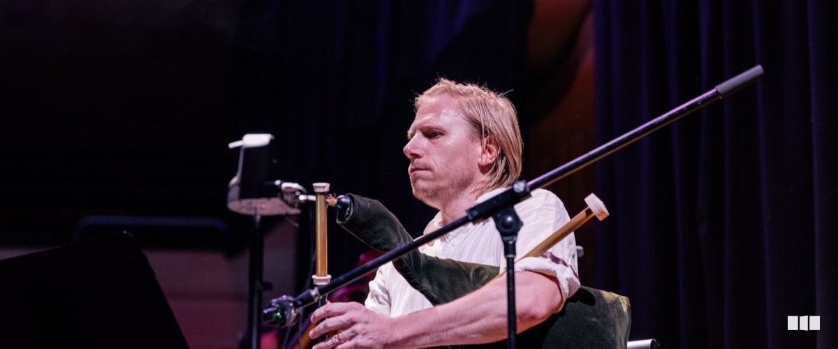 A male musician with shoulder-length blonde hair performs on bagpipes during an orchestral concert on a darkened stage.