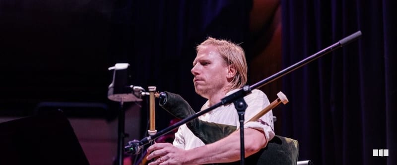 A male musician with shoulder-length blonde hair performs on bagpipes during an orchestral concert on a darkened stage.