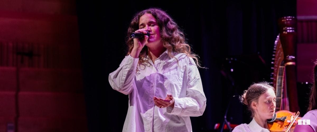 A female soloist in a white shirt sings into a handheld microphone on stage, with a harpist and violinist visible in the background.