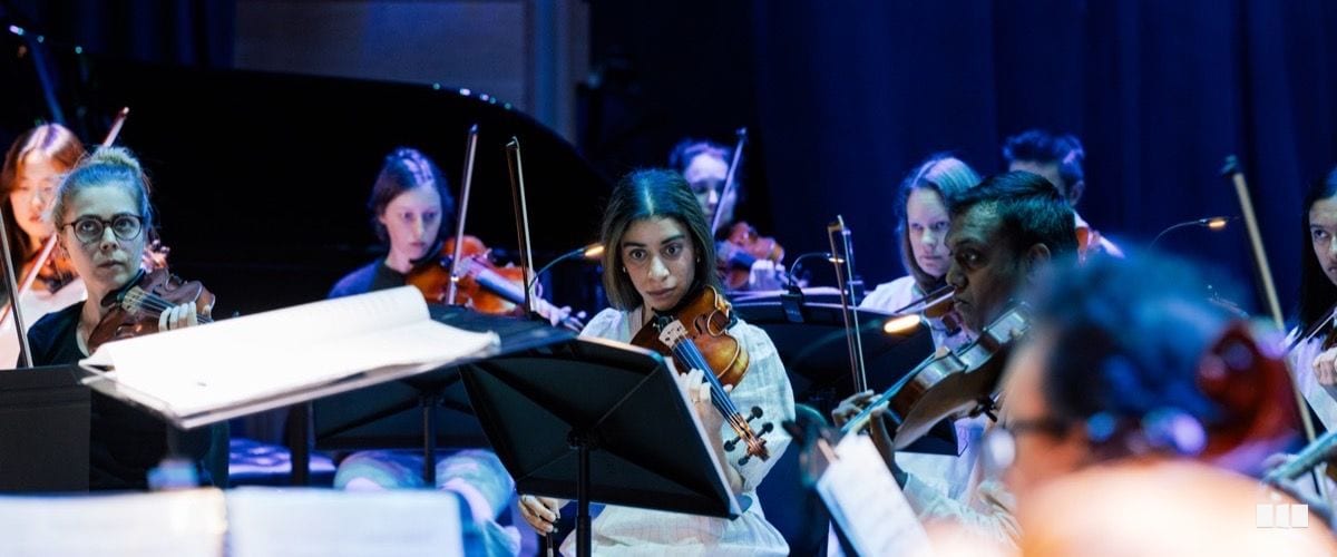 A diverse group of string players perform together in a dimly lit concert hall, reading from music stands.