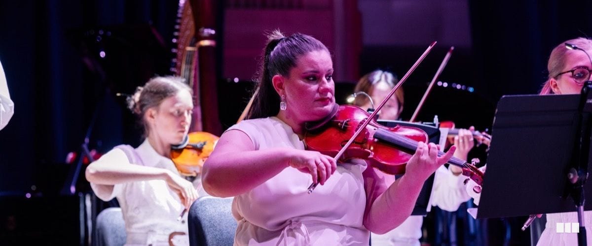 A violinist in a white dress performs with focused intensity alongside other string players in an orchestral concert setting.