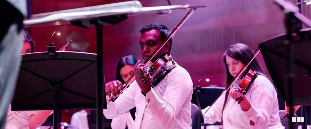 A violinist in a white shirt performs with focused intensity alongside fellow string players during an orchestral concert under warm stage lighting.