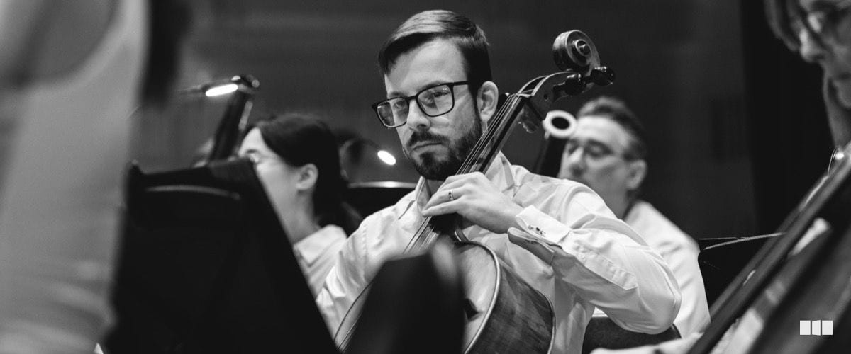 A bearded male cellist wearing glasses performs intently with fellow orchestra musicians visible in the background.