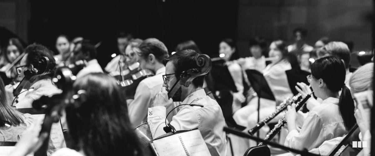 Orchestra musicians in white concert attire perform from sheet music on stands during a symphony concert.