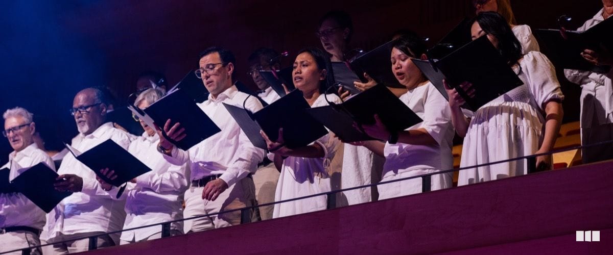 A choir dressed in white performs from an elevated balcony tier, holding black music folders during a concert.