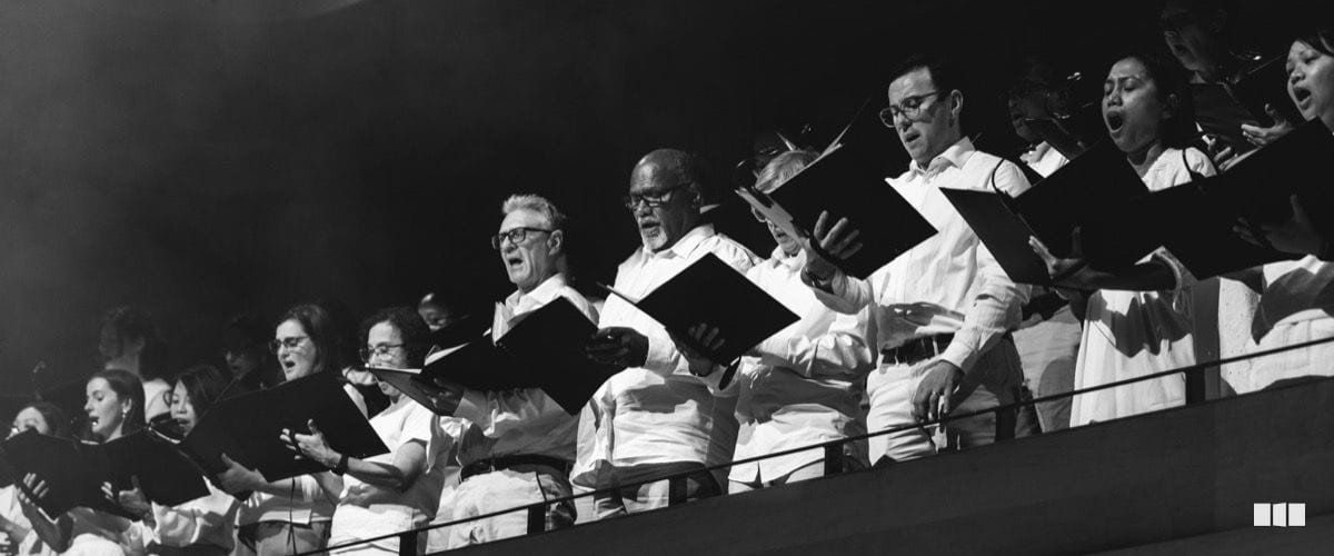 A choir performs from a balcony tier, holding black score folders and singing in white shirts against a dark background.