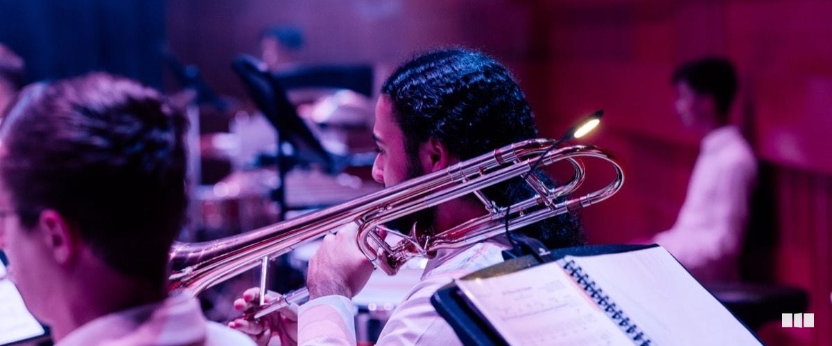 A trombone player performs in a concert setting, reading from sheet music under warm stage lighting.