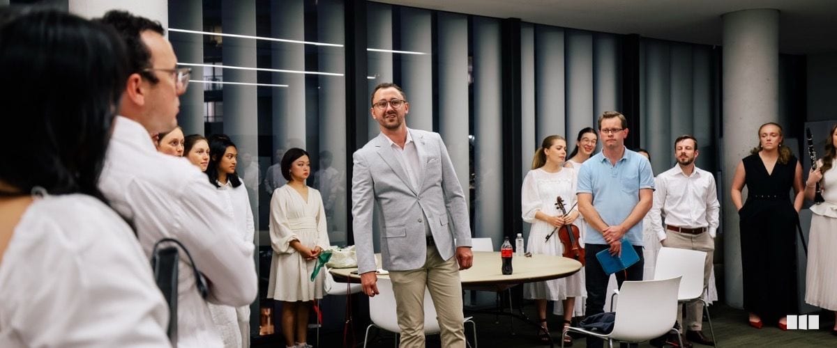 A man in a light grey blazer addresses a group of musicians and singers gathered in a modern rehearsal or reception space, with a violinist and clarinettist visible among the ensemble.