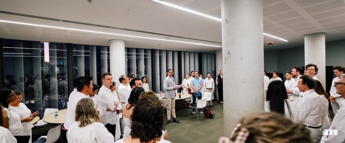 A large group of musicians dressed in white gather in a modern foyer space, listening to a speaker address the ensemble before or after a performance.