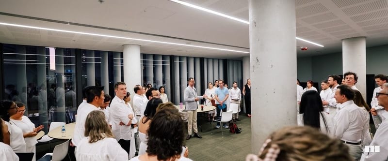 A large group of musicians dressed in white gather in a modern foyer space, listening to a speaker address the ensemble before or after a performance.