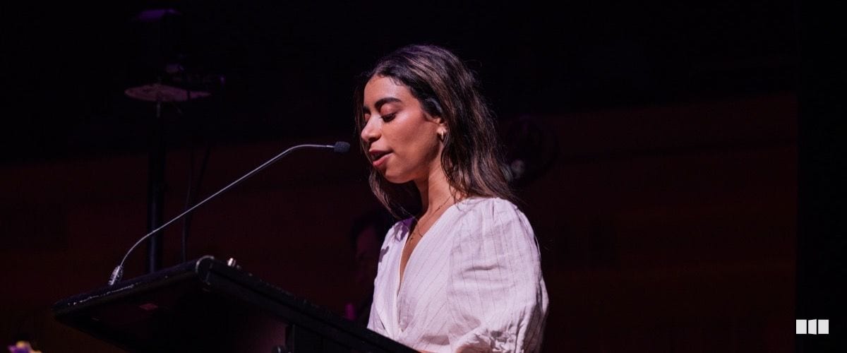 A young woman in a white blouse speaks at a podium microphone on a darkened stage