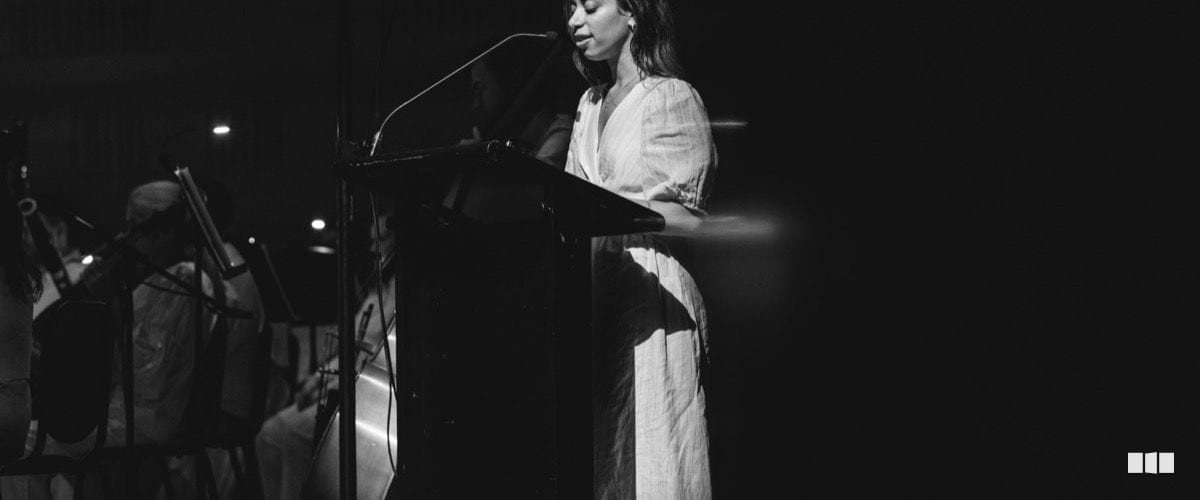 A woman in a white dress reads from notes at a podium on a darkened stage with musicians visible in the background.
