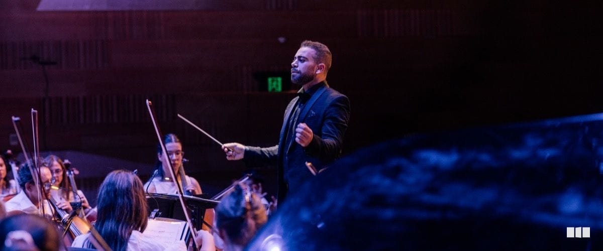 A conductor in a black suit leads an orchestra from the podium, baton raised, during a concert performance in a warmly lit concert hall.