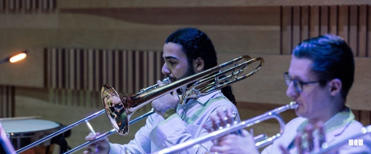 Two male trombonists perform together in a concert hall, both focused on playing their instruments