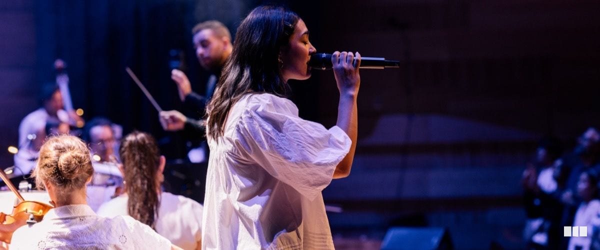 A female soloist in a white dress sings into a handheld microphone with an orchestra and conductor visible behind her on stage.