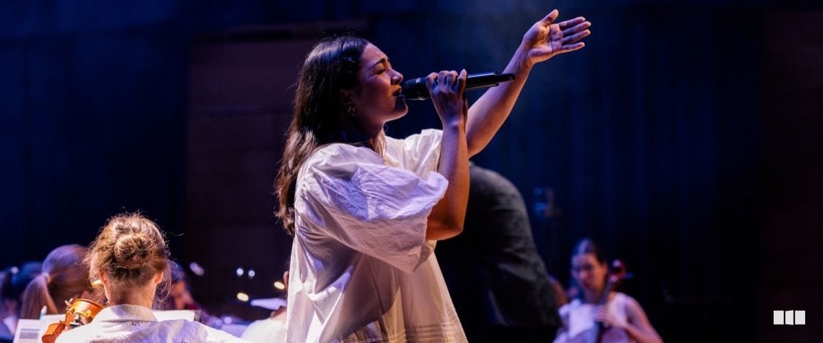 A female soloist in a flowing white dress sings passionately into a handheld microphone with one arm raised, backed by an orchestra on a dramatically lit stage.