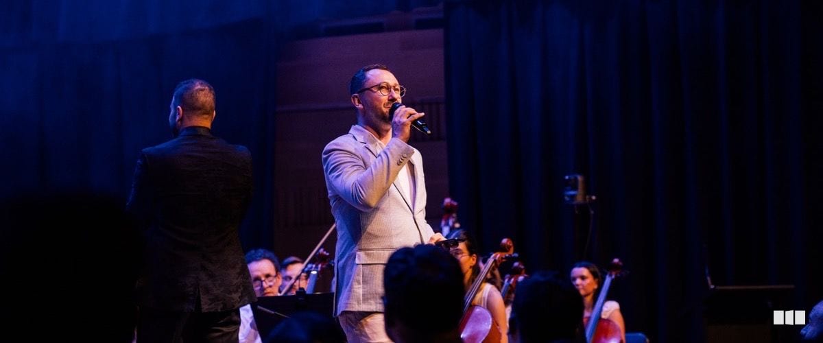 A male soloist in a light grey suit sings into a microphone on stage while an orchestra performs behind him and a conductor stands with his back to the camera