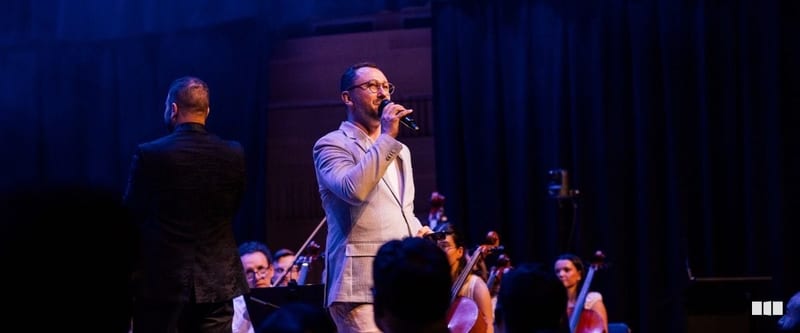 A male soloist in a light grey suit sings into a microphone on stage while an orchestra performs behind him and a conductor stands with his back to the camera