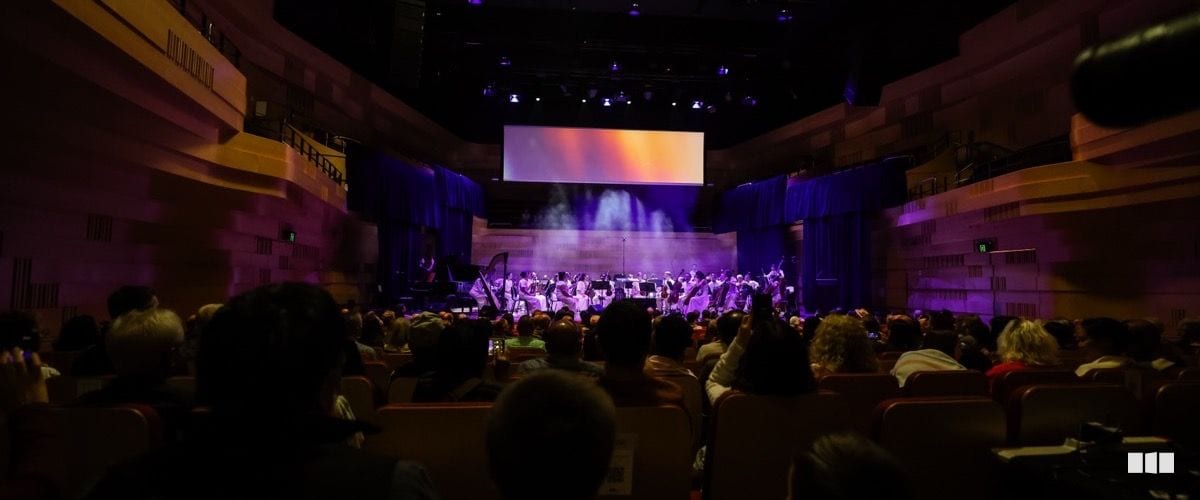 A symphony orchestra performs on a dramatically lit stage with purple lighting and a large projection screen before a full seated audience in a concert hall