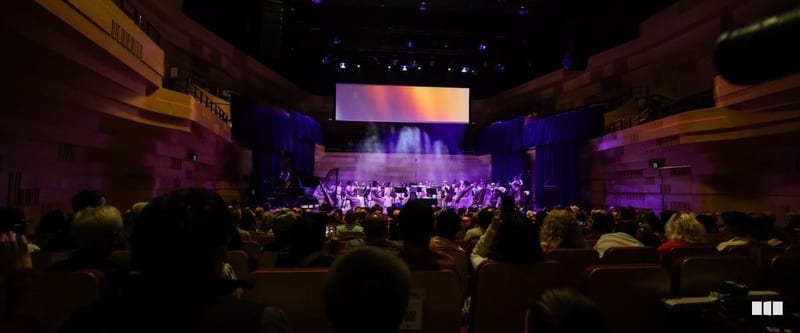 A symphony orchestra performs on a dramatically lit stage with purple lighting and a large projection screen before a full seated audience in a concert hall