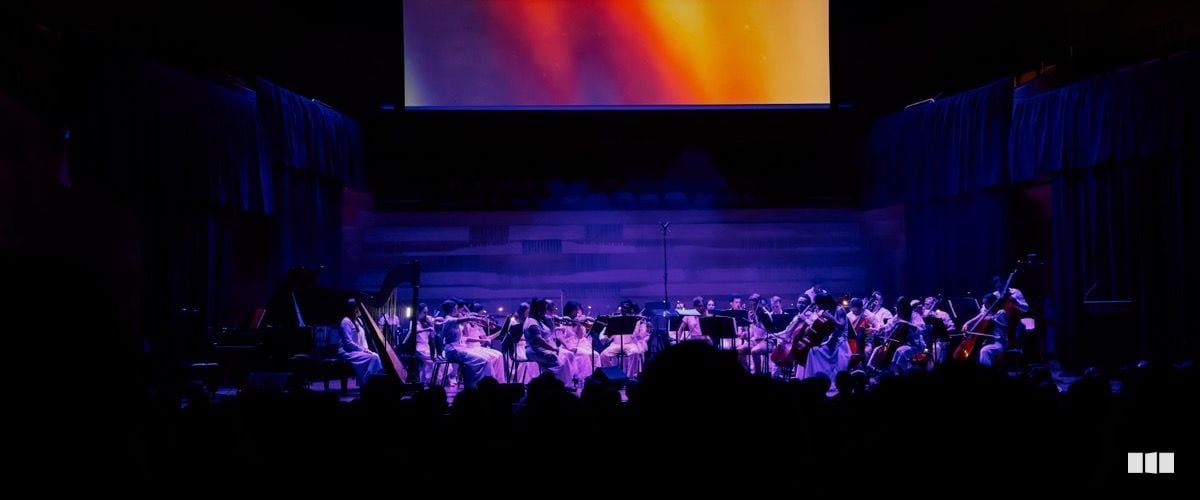 A full orchestra dressed in white performs on a dimly lit stage beneath a colourful projected screen.