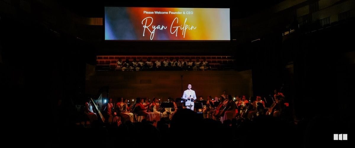 Founder and CEO Ryan Gilpin is welcomed on stage before a seated symphony orchestra with a choir visible on the balcony behind