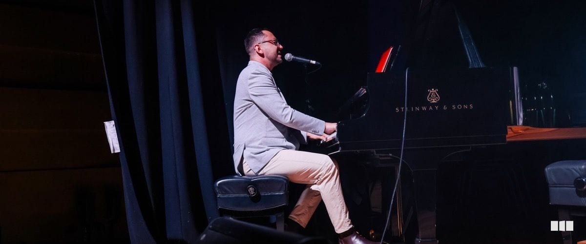 A male pianist performs and sings into a microphone at a Steinway & Sons grand piano on a darkened stage