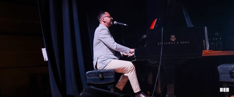 A male pianist performs and sings into a microphone at a Steinway & Sons grand piano on a darkened stage