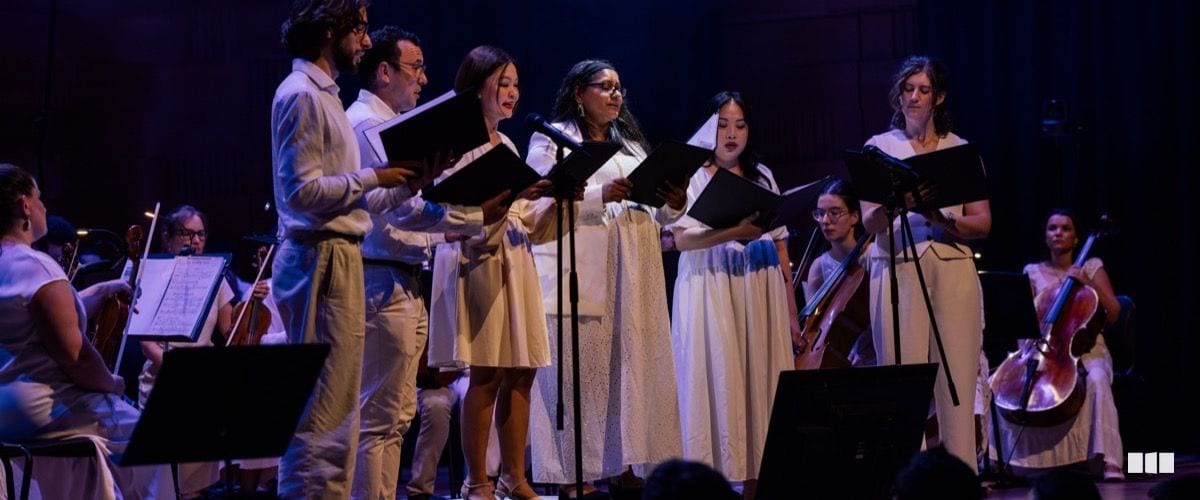 A small ensemble of soloists in white attire sing from black score folders on a dramatically lit stage, accompanied by a string orchestra seated behind them.