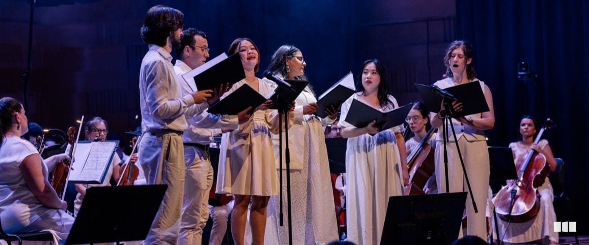 A group of choral soloists dressed in white perform on stage holding black score folders, accompanied by an orchestra in the background under dramatic blue stage lighting.