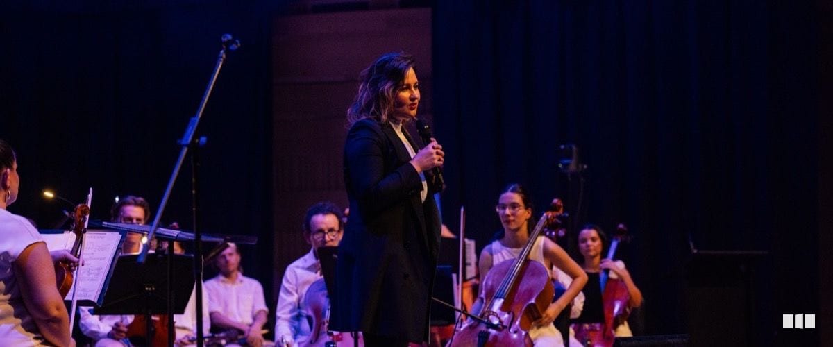 A woman in a black blazer speaks into a microphone on stage while string musicians sit behind her during an orchestral concert.