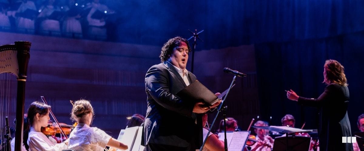 A male soloist sings from a score at a microphone stand while a conductor leads the orchestra behind him on a dramatically lit concert stage.