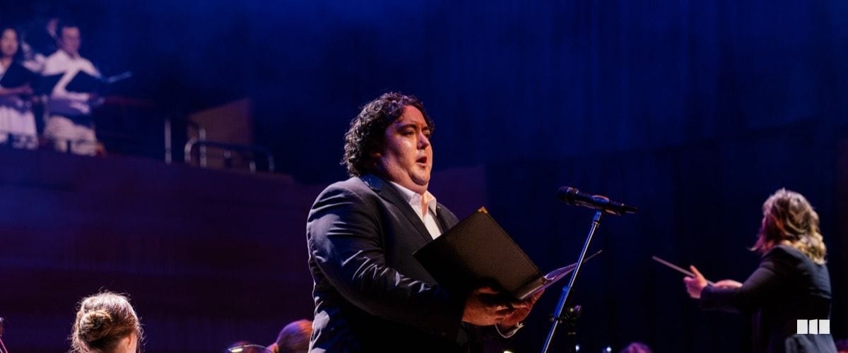 A male soloist holding a score folder sings into a microphone on a concert stage, with a conductor and choir visible in the background.