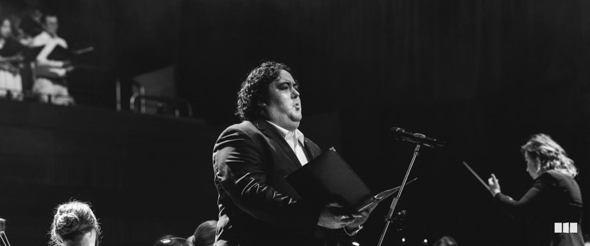 A suited soloist holds a score and sings into a microphone while a conductor leads the orchestra behind him.
