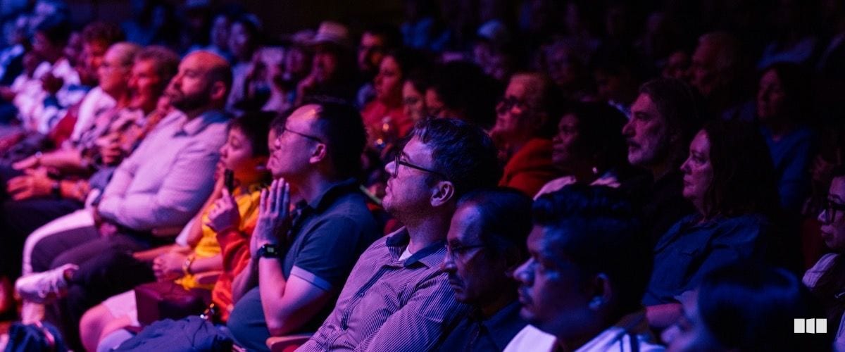 A diverse audience of adults sits attentively in a darkened concert hall illuminated by warm purple and red stage lighting.