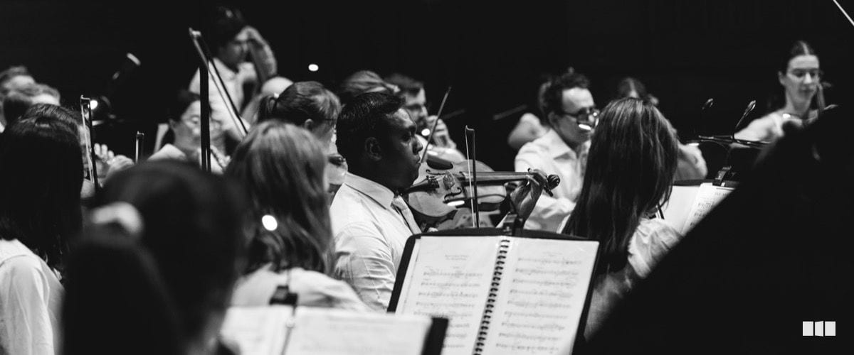 Orchestra musicians concentrate during a performance, with a violinist in focus amid a full ensemble and open sheet music in the foreground.