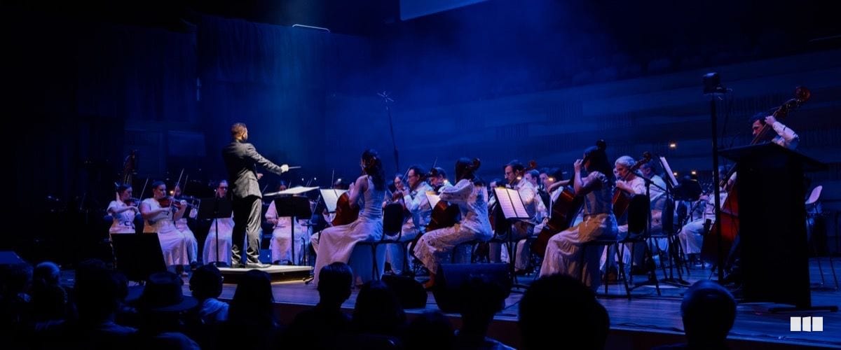 A conductor leads a full orchestra dressed in white under dramatic blue stage lighting during a concert performance.