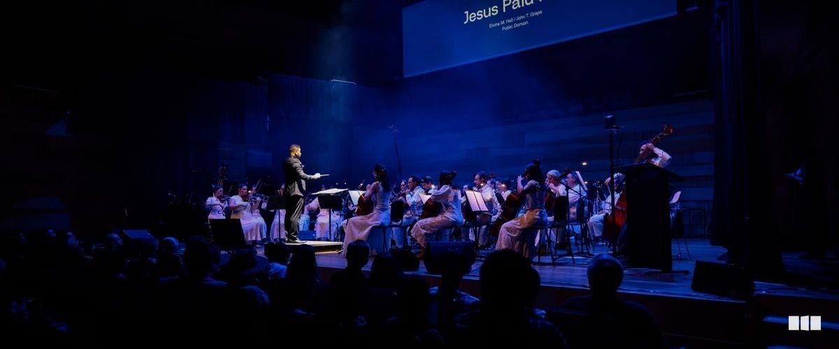 A conductor leads a full orchestra dressed in white and black formal attire on a blue-lit stage during a Christian concert performance.