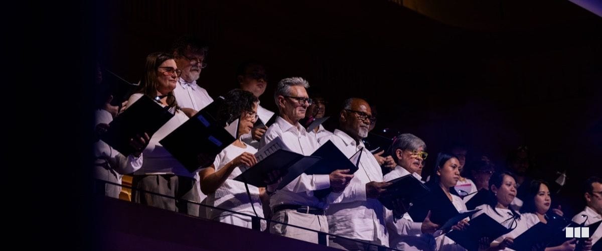 A diverse choir in white shirts holds music folders and sings from a balcony tier during a concert performance.