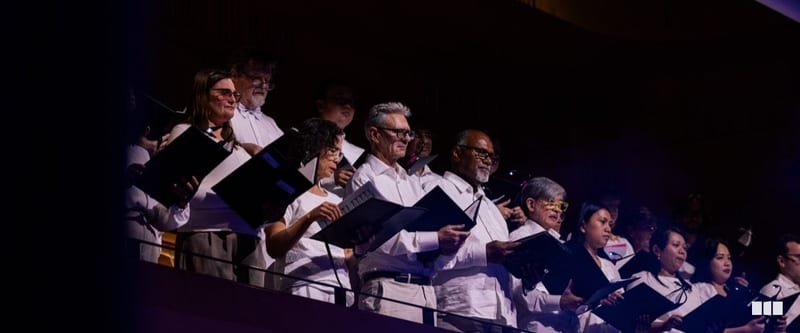 A diverse choir in white shirts holds music folders and sings from a balcony tier during a concert performance.