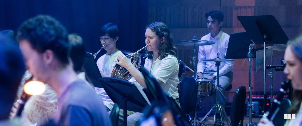 Student musicians play French horn, trumpet, clarinet, and drums during an orchestra rehearsal under blue stage lighting.