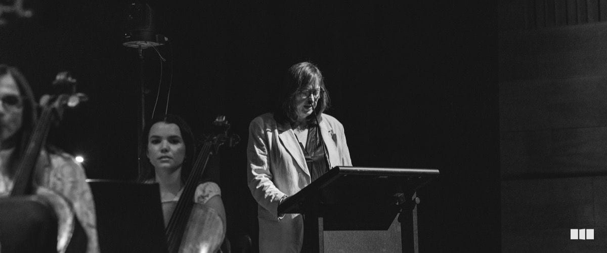 A woman in a light blazer speaks at a lectern on stage while string players are visible in the background.