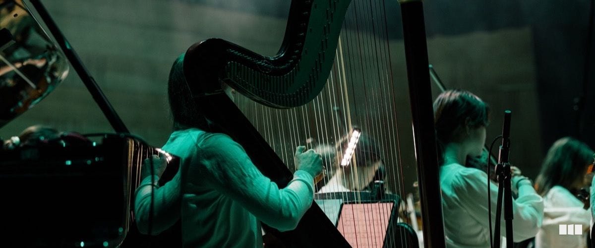 A harpist performs on stage under teal stage lighting during an orchestral concert, with other musicians visible in the background.
