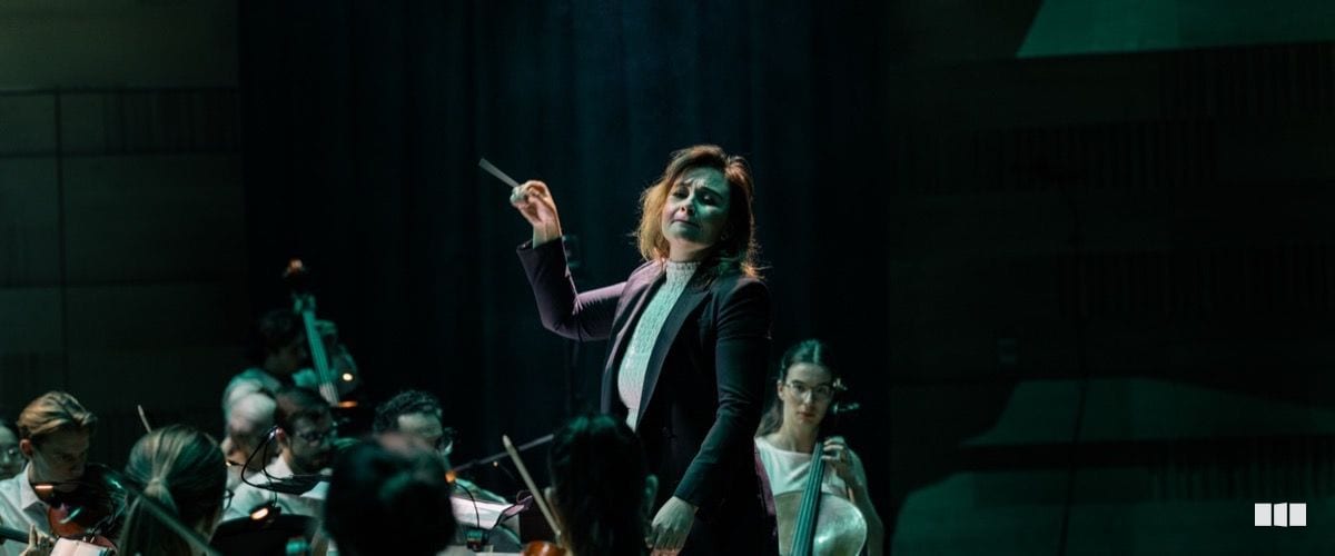 A female conductor raises her baton with expressive intensity while leading an orchestra on a darkly lit stage