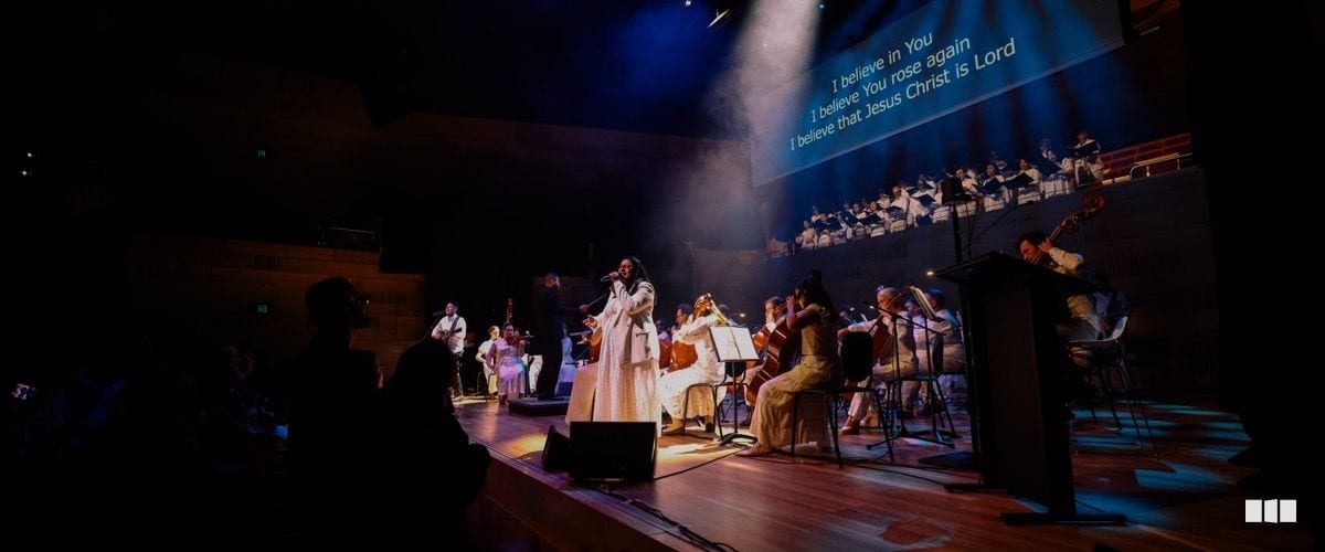 A soprano soloist in a white dress performs at centre stage with a full orchestra and choir during a Christian worship concert, lyrics projected on a screen behind.